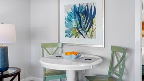 Un coin repas avec une table ronde blanche, deux chaises vertes, un saladier rempli d’oranges et une œuvre d’art encadrée accrochée au mur.
