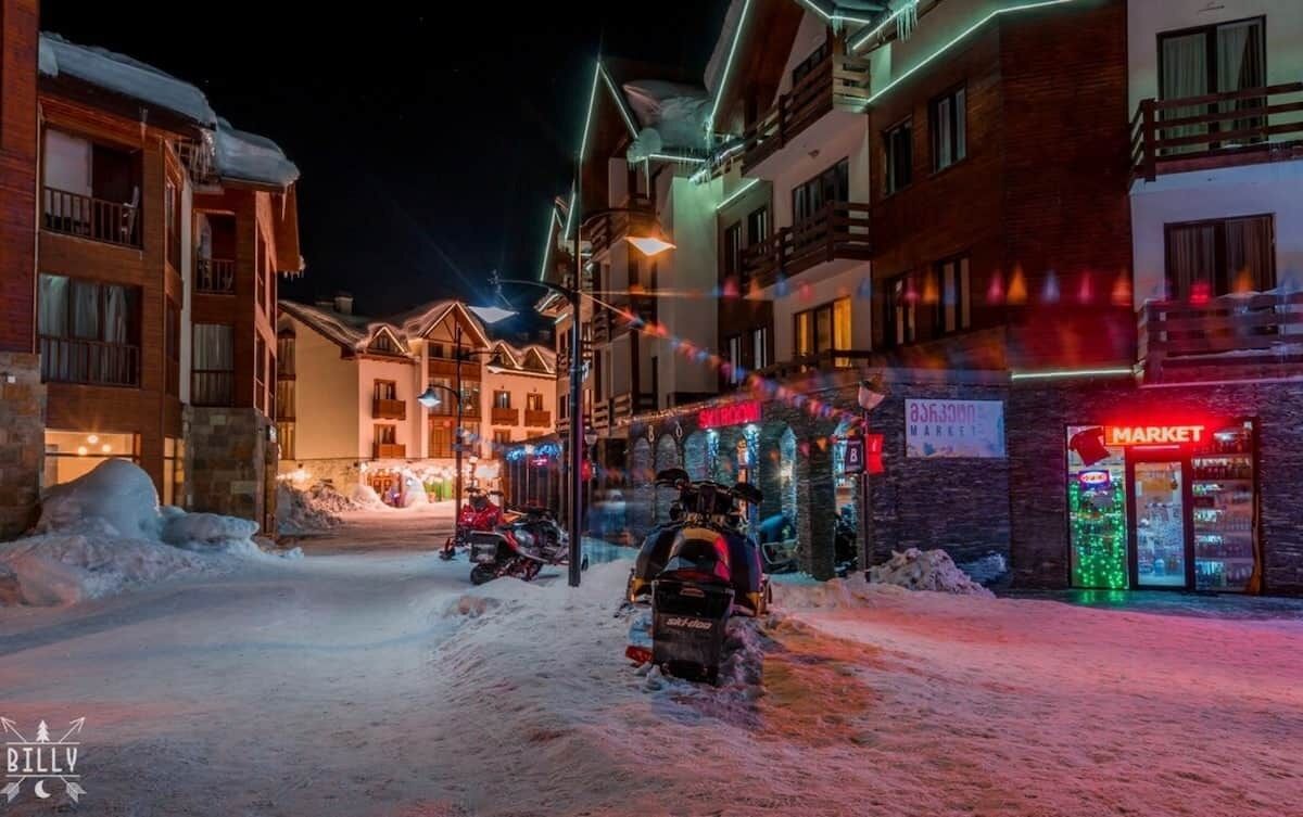 A snow-covered street with buildings, a market, and parked snowmobiles.