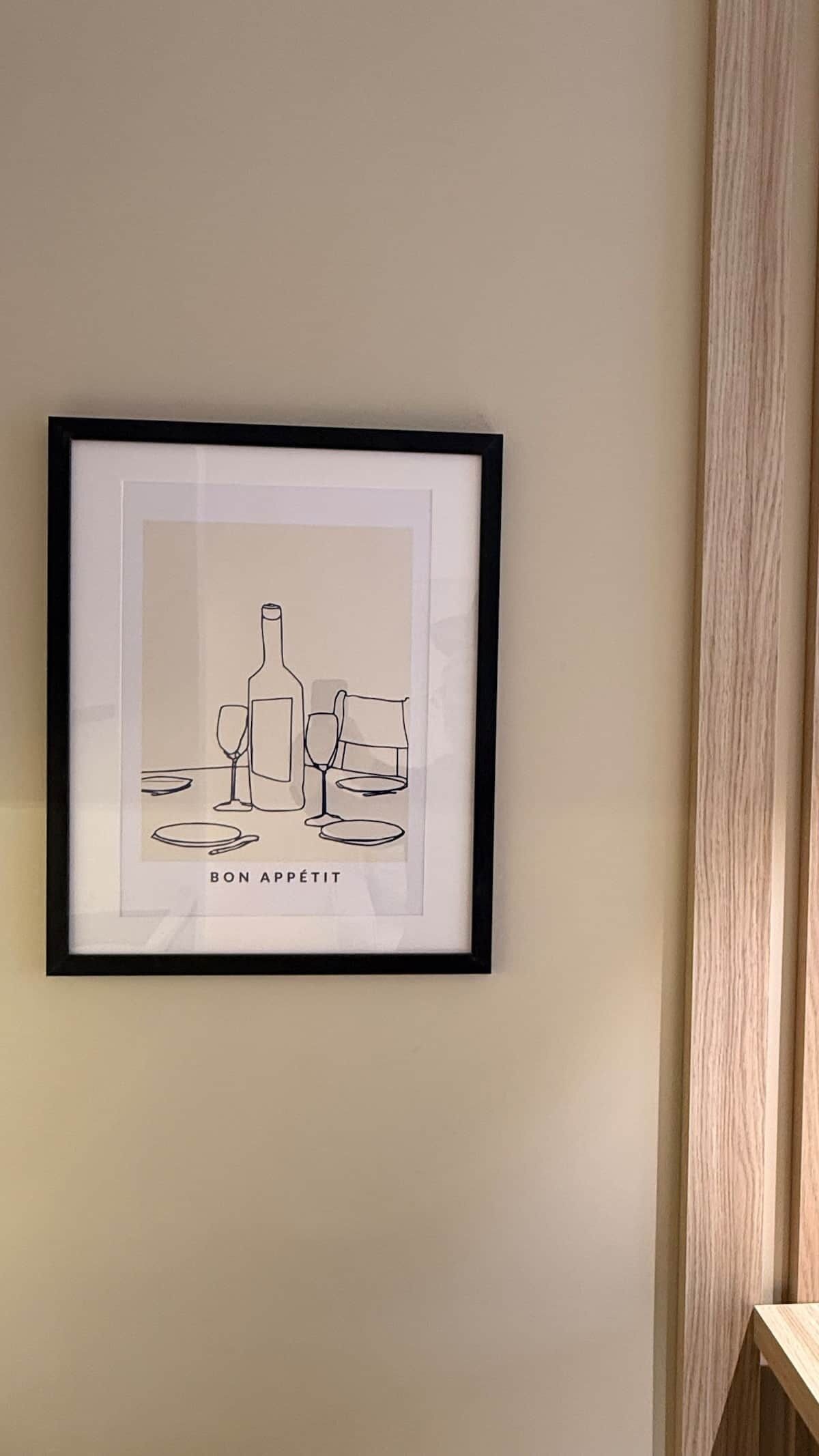 Ein gerahmtes Kunstwerk zeigt eine Weinflasche, Gläser und Teller, mit dem Schriftzug 'BON APPETIT' an einer Wand.