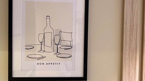 Ein gerahmtes Kunstwerk zeigt eine Weinflasche, Gläser und Teller, mit dem Schriftzug 'BON APPETIT' an einer Wand.