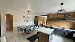 Uma cozinha moderna com uma ilha central, uma sala de jantar com uma mesa grande e cadeiras, e uma porta visível que dá para outro cômodo.