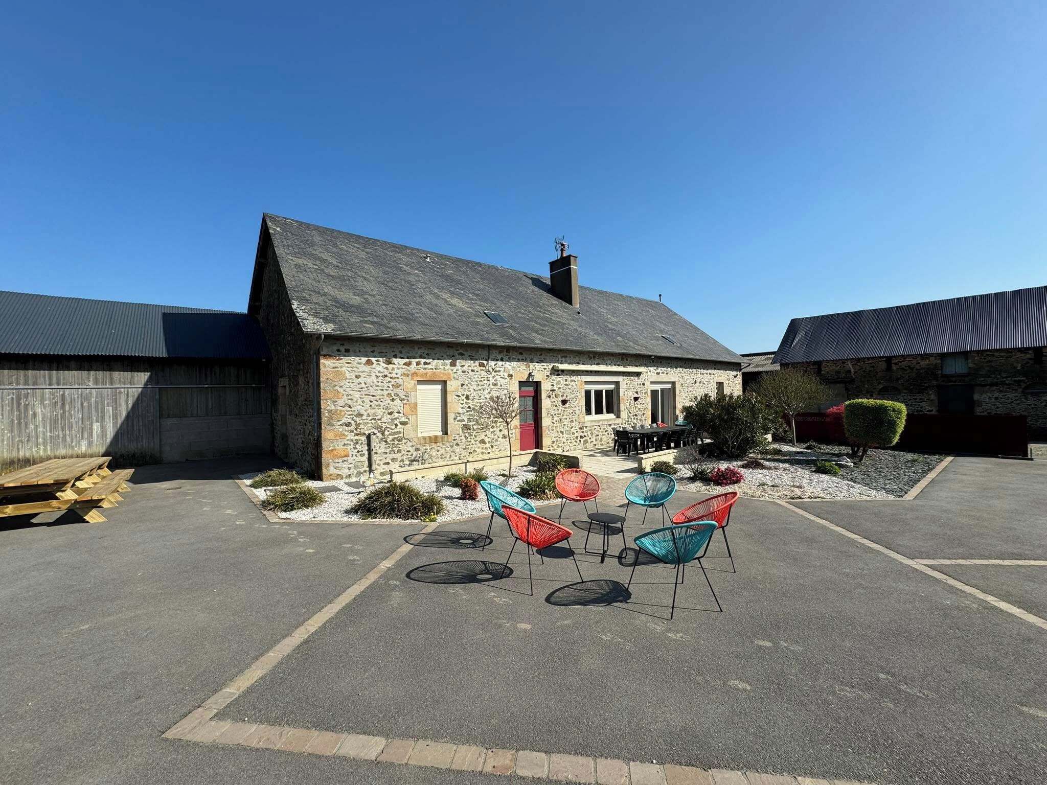 A stone building with a red door and a chimney, a covered outdoor seating area with colorful chairs, and a paved parking lot.