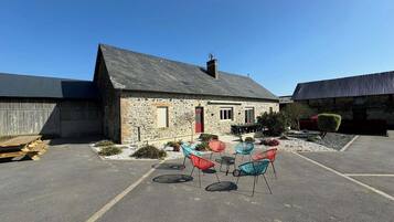 A stone building with a red door and a chimney, a covered outdoor seating area with colorful chairs, and a paved parking lot.