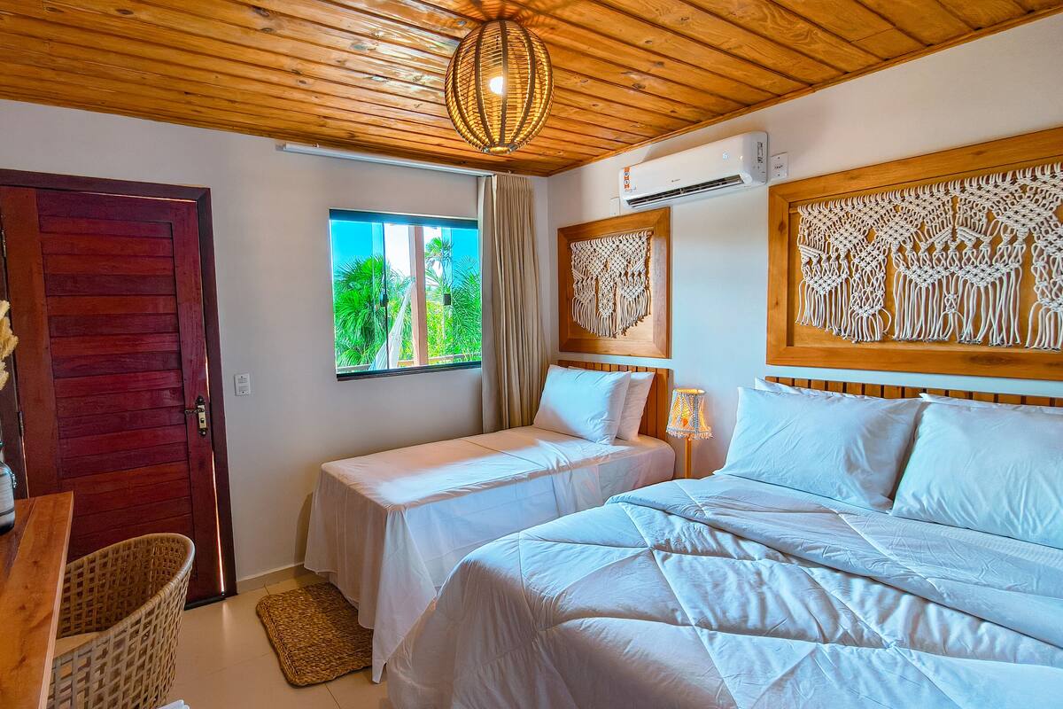 Una habitación con techo de madera, cama con ropa de cama blanca, ventana con vistas a la vegetación y un tapiz de macramé en la pared.