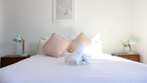 Een opgemaakt bed met opgerolde handdoeken, twee nachtkastjes met lampen, en een ingelijst schilderij aan de muur.