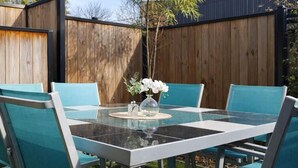 Een tafel met glazen blad, stoelen, een bloemenvaas en een houten hek op de achtergrond.