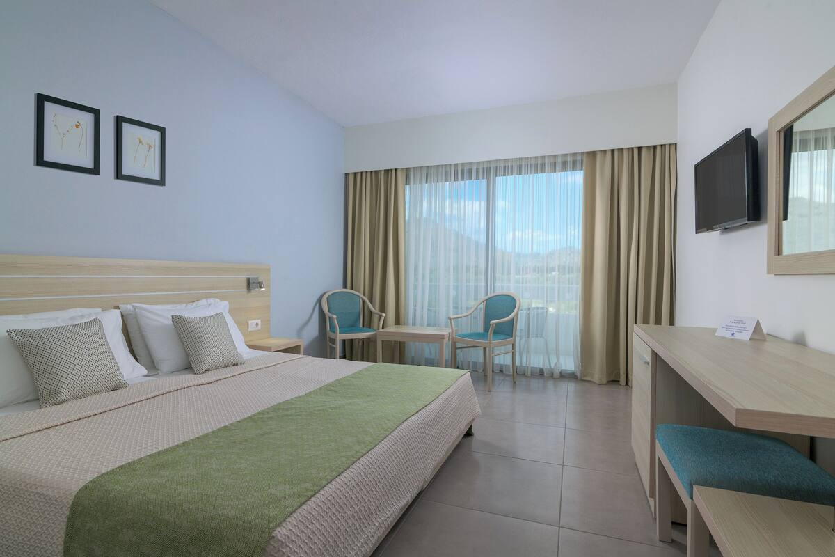 Una camera d'albergo con un letto, una scrivania, due sedie, una TV e una finestra con tende.