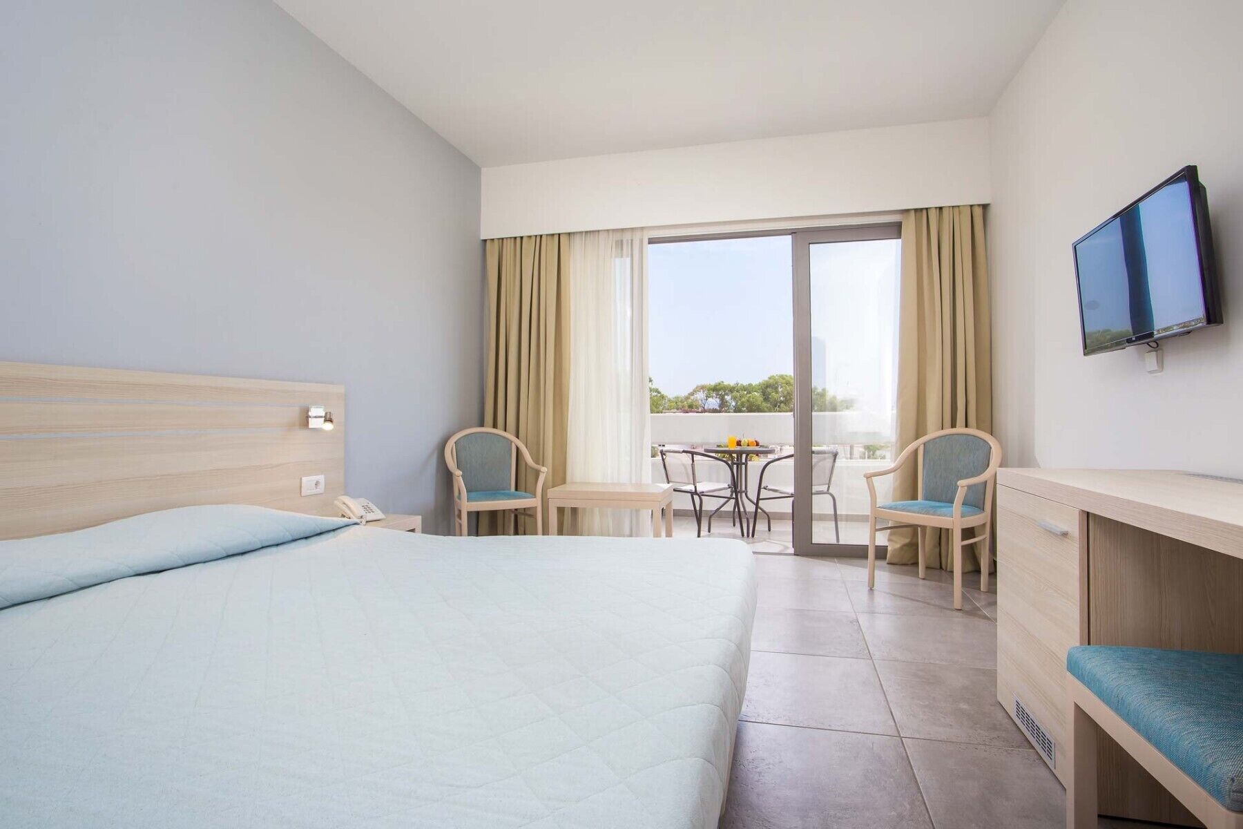 A hotel room with a bed, a desk, a chair, a TV, and a balcony with a table and chairs.