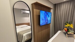 A hotel room with a bed, a TV mounted on a wooden panel, a mirror, and a small table with fruits and a cup. - Apartment steps away from the beach, ideal to enjoy the Brazilian Caribbean. (João Pessoa)