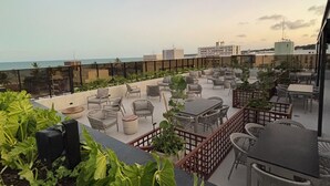 A rooftop terrace with outdoor seating, a view of the sea, and some greenery in the foreground. - Apartment steps away from the beach, ideal to enjoy the Brazilian Caribbean. (João Pessoa)