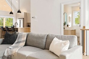 A modern living room with a grey sofa, a coffee table, and a dining area with chairs and a kitchen in the background. - Spa - River - Game Room | Le Belvedere (Mont-Tremblant)
