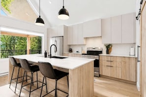 A modern kitchen with a central island, bar stools, stainless steel appliances, and a view of greenery outside. - Spa - River - Game Room | Le Belvedere (Mont-Tremblant)