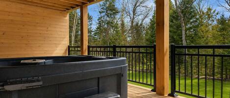 Un espacio al aire libre techado con jacuzzi, terraza de madera y vistas a un jardín con césped y árboles.