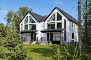 Ein modernes, zweistöckiges Haus mit großen Fenstern und einem schwarzen Dach, umgeben von Bäumen und Gras.