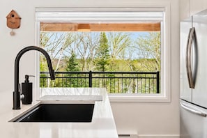 Une cuisine moderne avec un réfrigérateur en acier inoxydable, un robinet noir et une fenêtre donnant sur des arbres, ainsi qu’une balustrade de balcon.