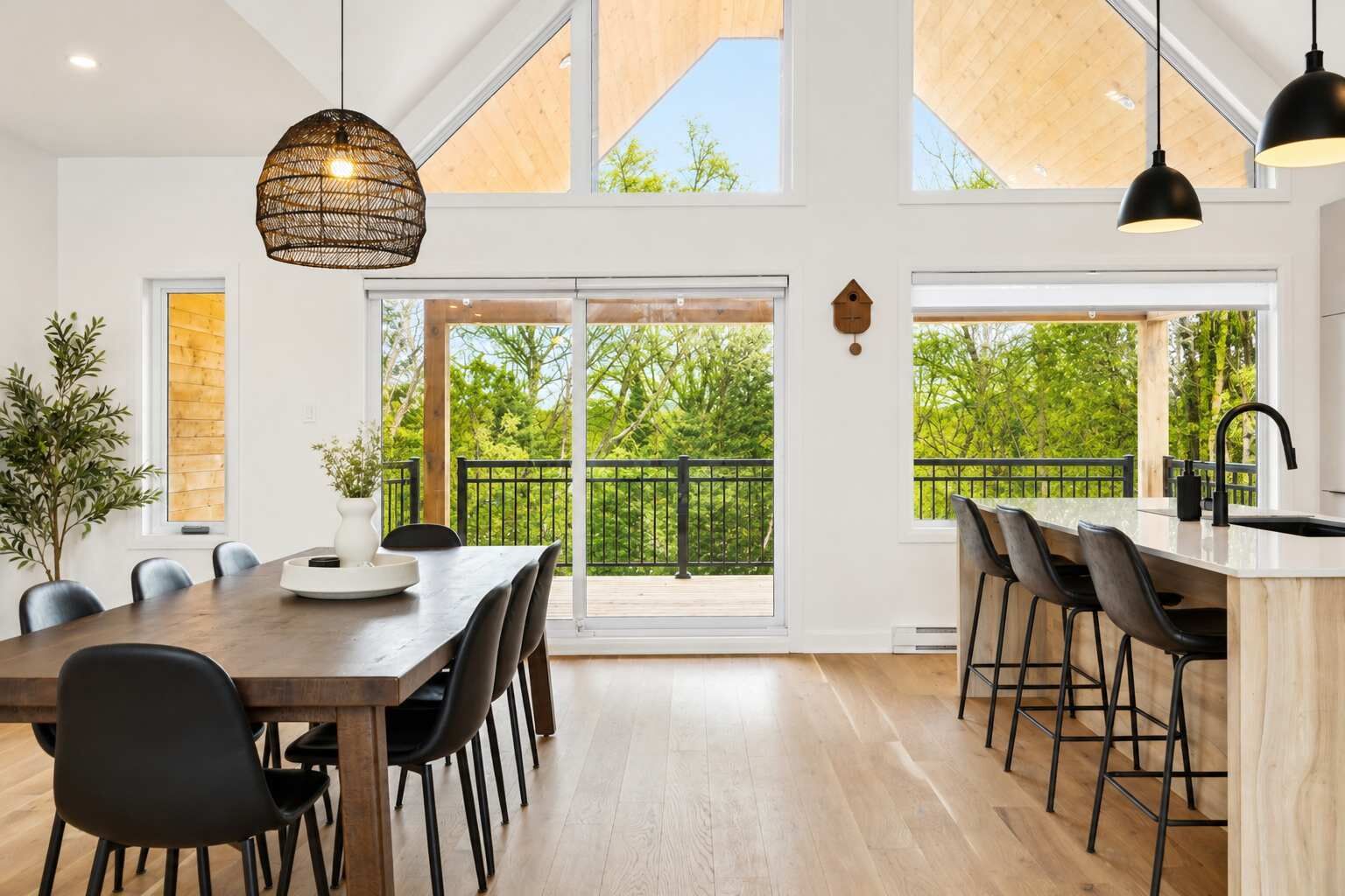 A modern kitchen with a large dining table, bar stools, and a view of the outdoors through large windows.