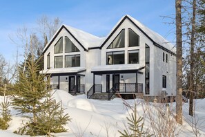A modern two-story house with large windows and a snow-covered roof, surrounded by trees. - Spa - River - Game Room | Le Boréal (Mont-Tremblant)