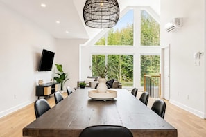 A modern dining room with a long table, chairs, a television, and a large window with a view of greenery. - Spa - River - Game Room | Le Boréal (Mont-Tremblant)
