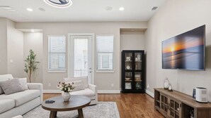Un salon moderne avec un canapé, une table basse et un téléviseur. Il y a trois fenêtres équipées de stores, un meuble et une plante.