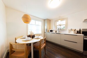 Une cuisine moderne avec une table de salle à manger ronde, des chaises en bois, un luminaire suspendu, un four encastré et une fenêtre laissant entrer la lumière naturelle.