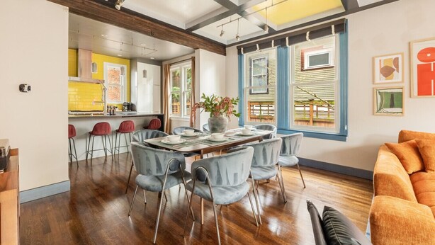 A spacious living area with a dining table set for four, a kitchen in the background, and large windows with blue frames. - NEW The Cap Hill Heartbeat | Walk to Shops & Cafés (Seattle)