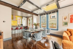 A spacious living area with a dining table set for four, a kitchen in the background, and large windows with blue frames.
