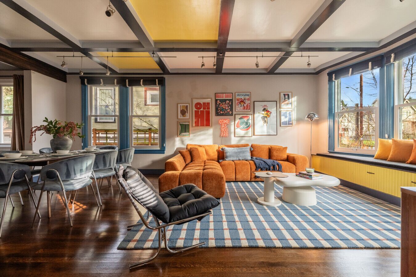A modern living room with a sofa, coffee table, and chairs. There are large windows with curtains, a dining table with chairs, and artwork on the walls.