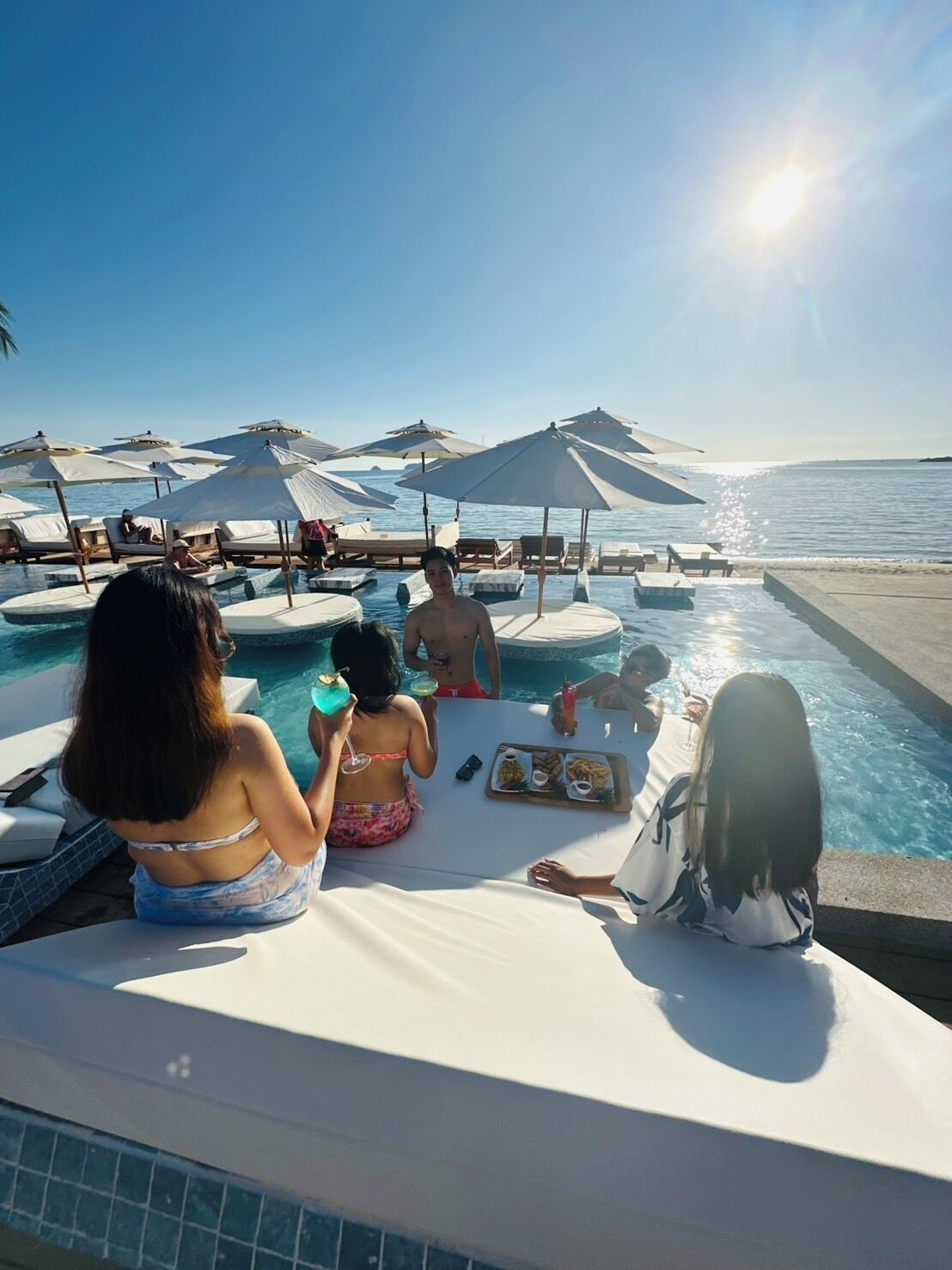 Ein Poolbereich mit Liegestühlen und Sonnenschirmen, ein Esstisch mit Speisen und Getränken und freie Sicht auf den Ozean.