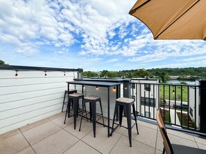 Eine Dachterrasse mit Barbereich, drei Barhocker und Blick auf einen Fluss und Grünflächen.