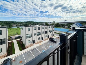 A multi-story building with a parking lot, a pedestrian pathway, and a view of a bridge and greenery in the background.