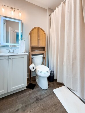 A bathroom with a white toilet, a wooden cabinet, a mirror with lights, and a shower curtain.