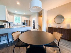 A modern kitchen with white cabinets, a round dining table with four chairs, a hanging light fixture, and a round mirror on the wall.