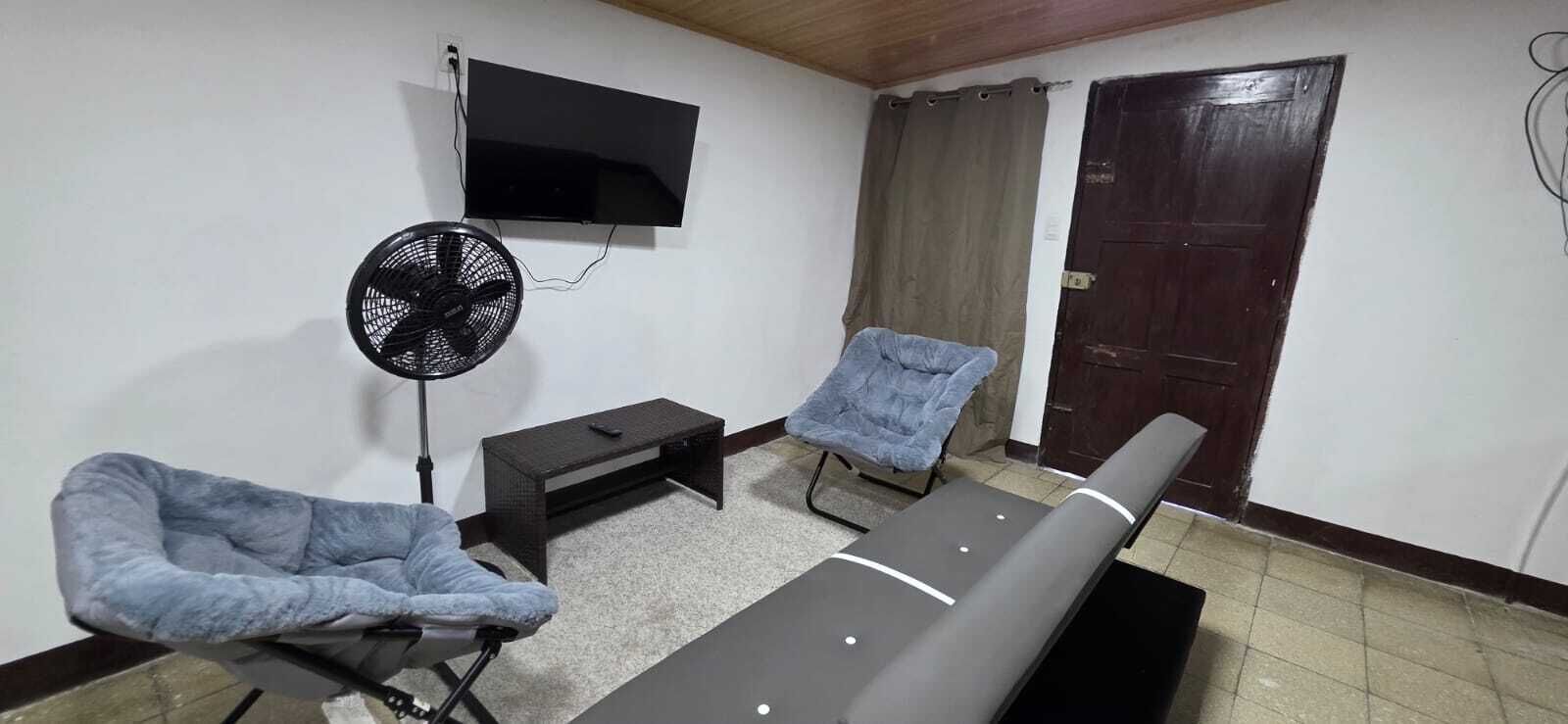 Une pièce comprenant un lit, une chaise, une petite table, un ventilateur et un téléviseur fixé au mur.