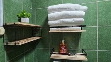 Un baño con azulejos verdes, un estante de madera con una planta en maceta, una pila de toallas blancas y un dispensador de jabón en una pequeña repisa.