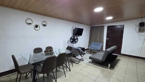 A room with a glass-top table, six chairs, a sofa, a standing fan, and a TV mounted on the wall.