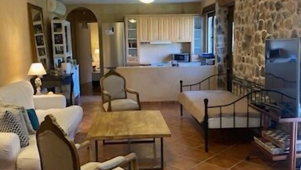 Un salon comprenant un canapé, deux fauteuils, une table basse et un téléviseur. Au fond, on aperçoit une cuisine avec des placards et un four à micro-ondes.