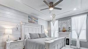 Un dormitorio con cama, mesitas de noche, cómoda, ventilador de techo y chimenea.