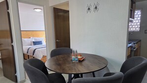 A hotel room with a round wooden table, four grey chairs, a bed with white bedding, a small kitchen area, and a window with a view. - Lofts by Verticca Héroes (Tecamac)