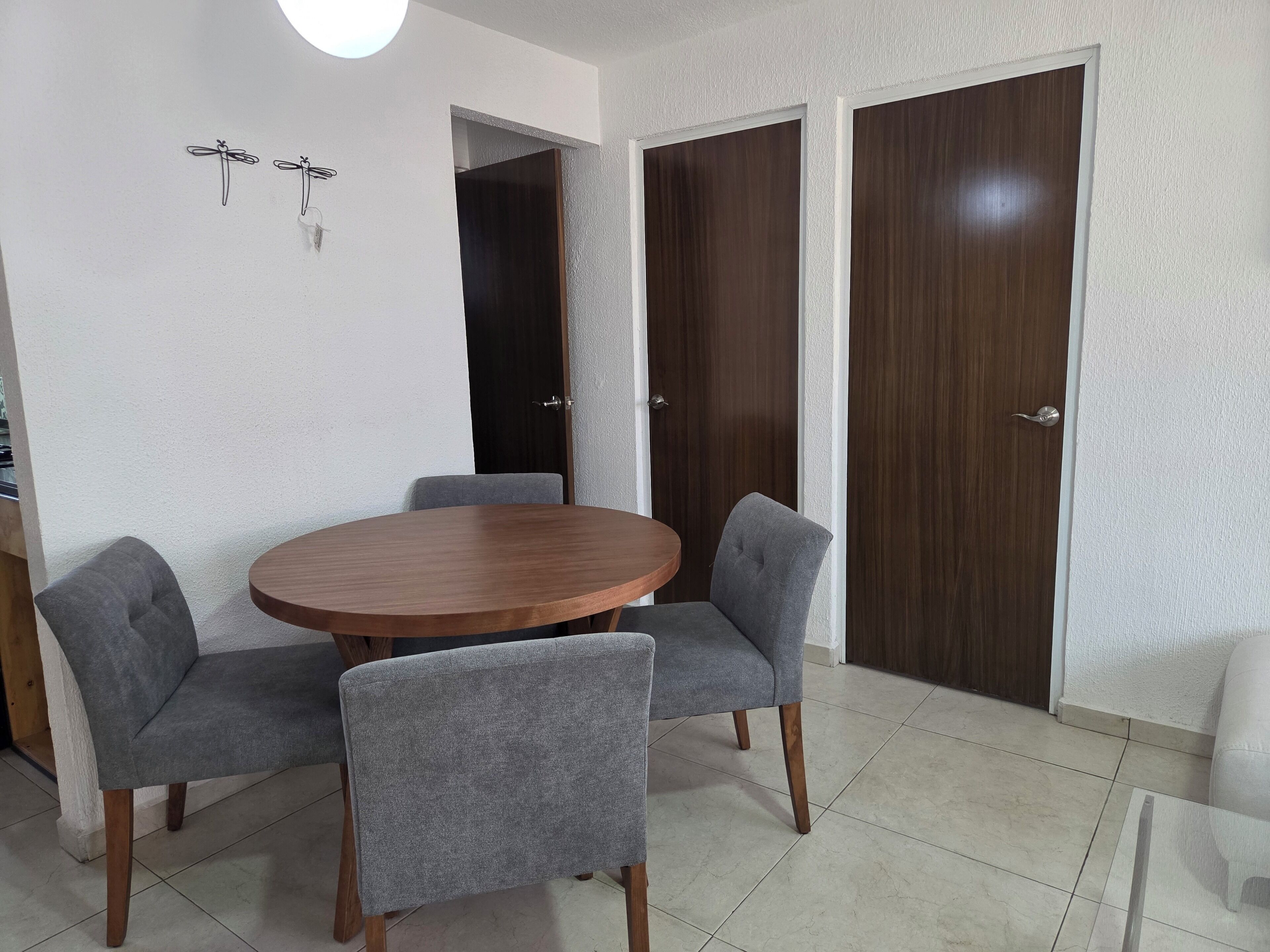Een eetruimte met een ronde houten tafel en vier grijs beklede stoelen. Twee donkerhouten deuren en een witte muur met een lamp.