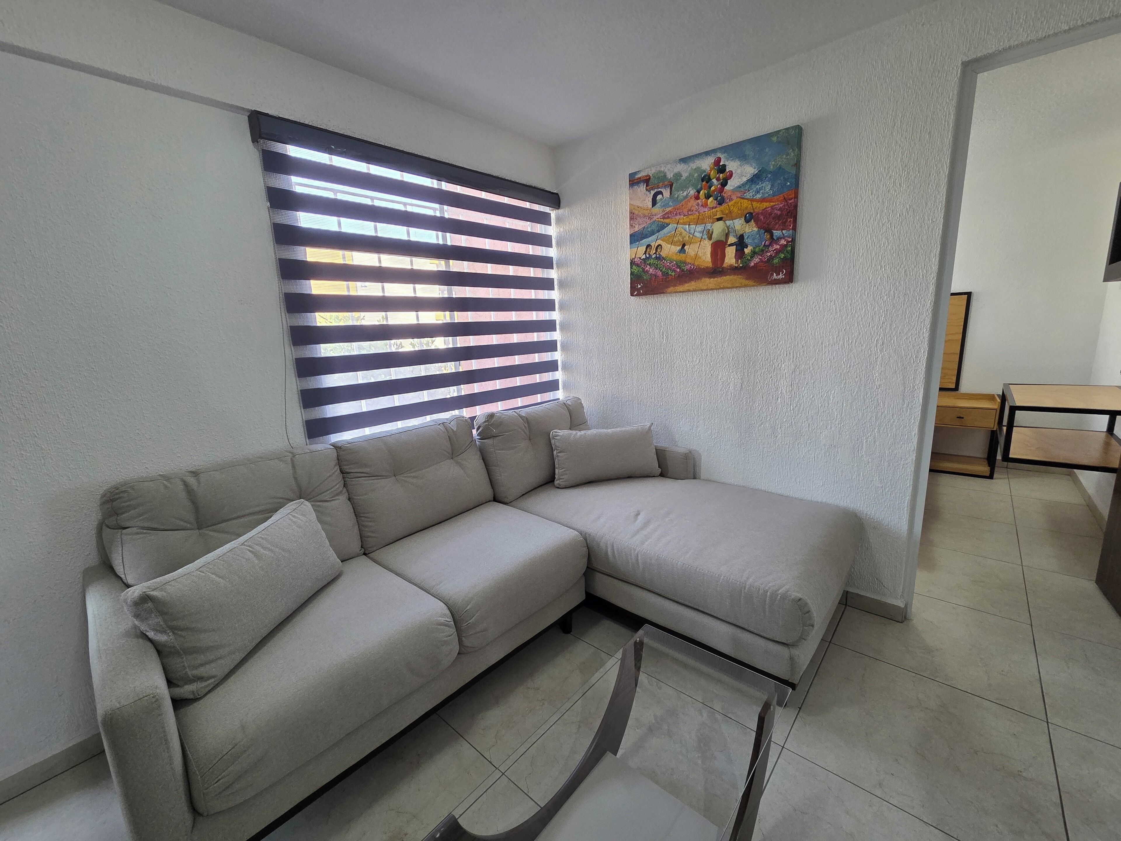 Una sala de estar con un sofá seccional color beige, una mesa de centro de vidrio, un cuadro en la pared y una ventana con persianas.