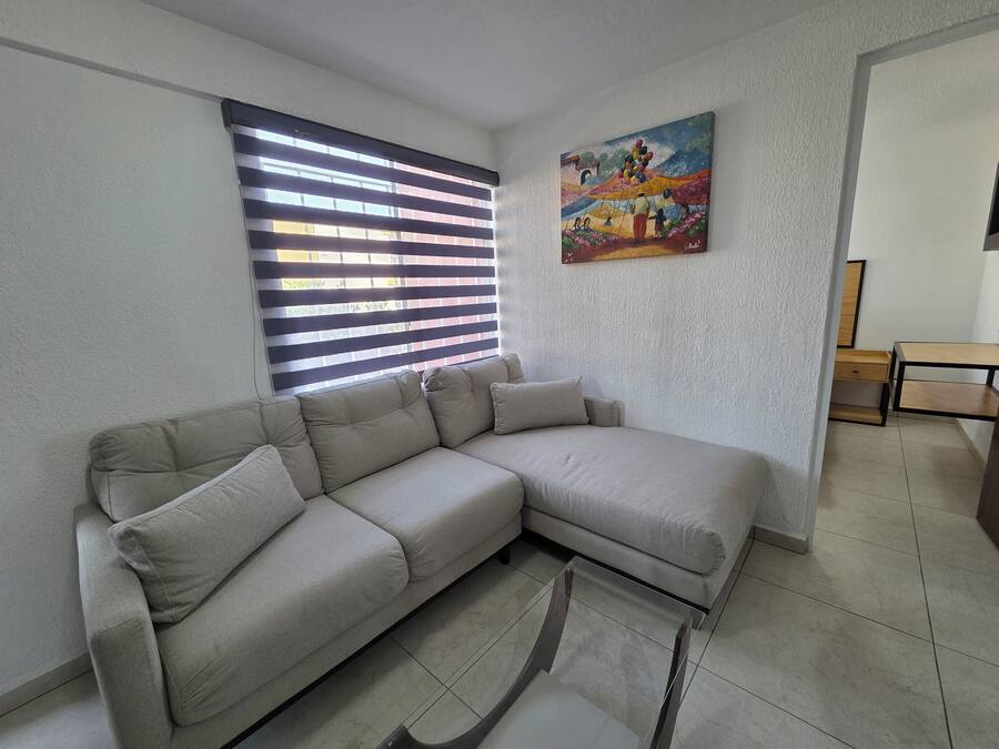 Una sala de estar con un sofá seccional color beige, una mesa de centro de vidrio, un cuadro en la pared y una ventana con persianas.