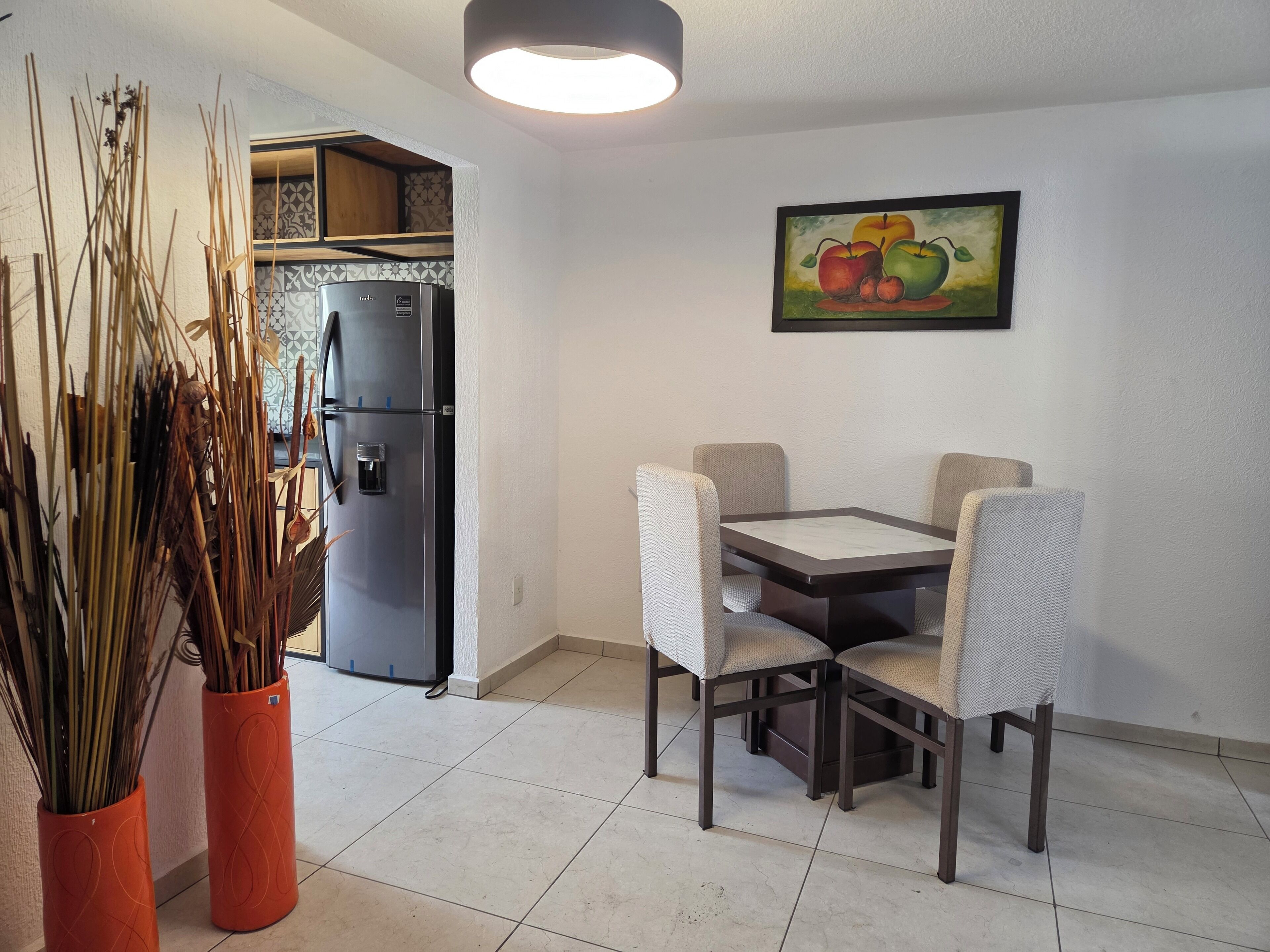 A compact kitchen with a refrigerator, a dining table with four chairs, and a painting of fruits on the wall.