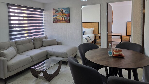 A modern living room with a sofa, a glass coffee table, a dining table with chairs, a painting on the wall, and a ceiling light. - Lofts by Verticca Héroes (Tecamac)