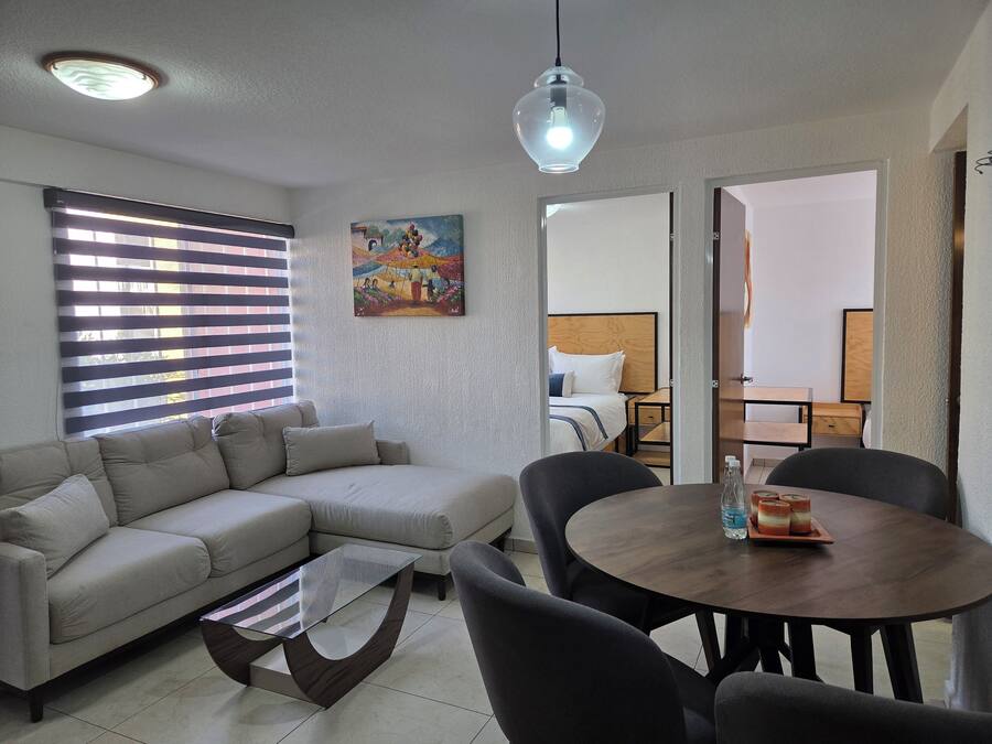 A modern living room with a sofa, a glass coffee table, a dining table with chairs, a painting on the wall, and a ceiling light.