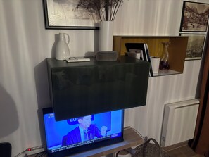 Ein modernes Wohnzimmer mit einem Flachbildfernseher, der ein Nachrichteninterview zeigt, einer Kaffeemaschine, einer Fernbedienung und Dekorationsobjekten auf einem Glastisch.