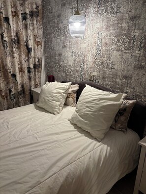 Ein Bett mit weißer Bettwäsche und mehreren Kissen, ein Nachttisch mit einem roten Gegenstand und eine strukturierte Wand mit einer hängenden Leuchte.