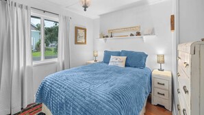 Een slaapkamer met een groot bed, een raam met gordijnen, een nachtkastje met een lamp en een dressoir.