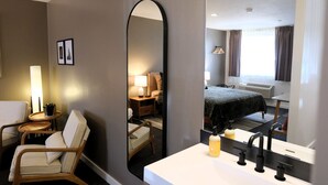 Ein Hotelzimmer mit einem großen Spiegel, einem Bett mit dunkler Bettdecke, einem Nachttisch mit Lampe, einem Sessel und einem kleinen Tisch mit Pflanze.
