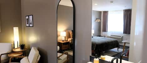 Habitación de hotel con un espejo grande, una cama con cubrecama oscuro, una mesita de noche con lámpara, una silla y una pequeña mesa con una planta.