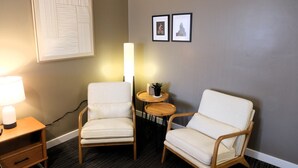 Ein Zimmer mit zwei weißen Sesseln, einem kleinen runden Tisch mit einer Pflanze, einer Wandlampe und gerahmten Bildern.